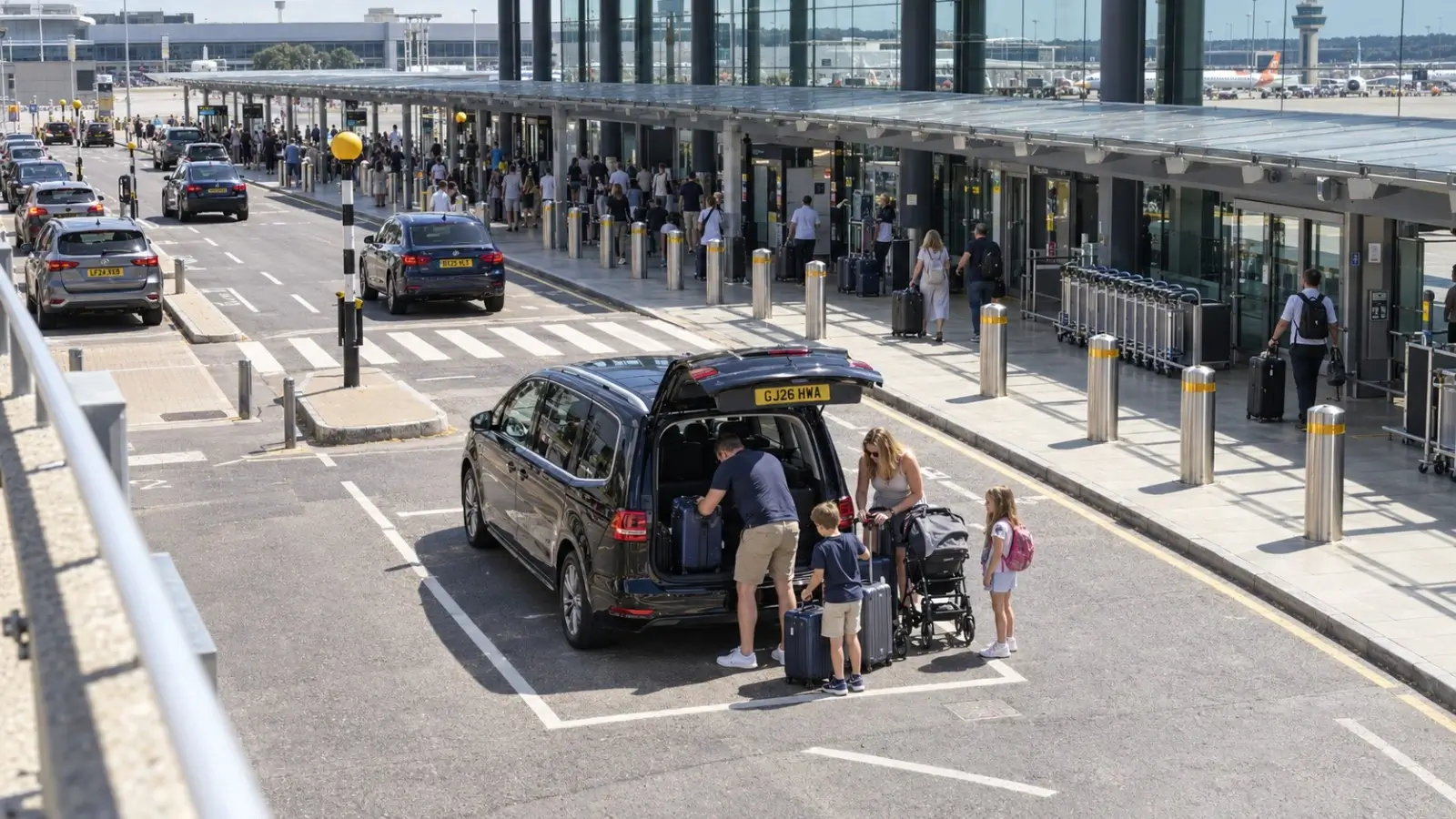 Black VW Sharan six-seater taxi loading family luggage at Gatwick South Terminal in midday sun